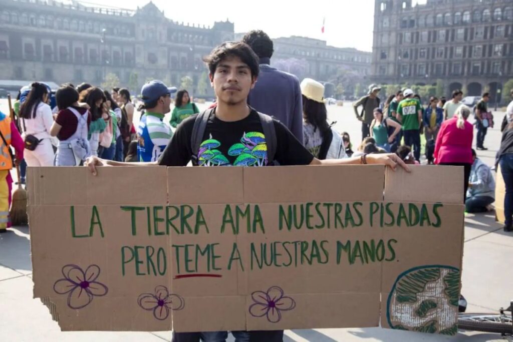 Joven sosteniendo cartel con la frase "La tierra ama nuestras pisadas, pero teme a nuestras manos" en una protesta ambiental.