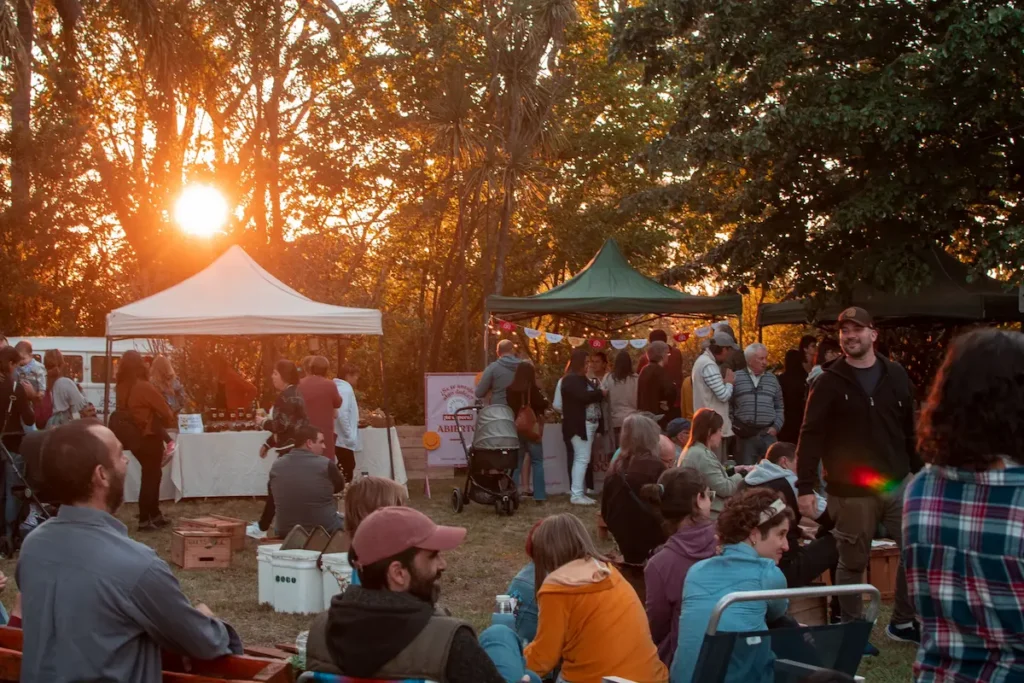 Feria Ecológica de Colonia Valdense, Uruguay, mostrando la comunidad sentada en el pasto, con stands de exposición, carpas y árboles al atardecer.
