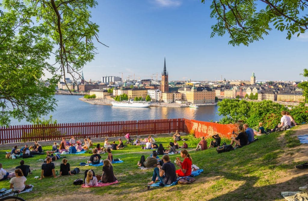 Young people spend time (sit, drink, eat) on lawn with green grass and trees around on Sodermalm island with view of old historical town Riddarholmen and Gamla Stan | Ambienta