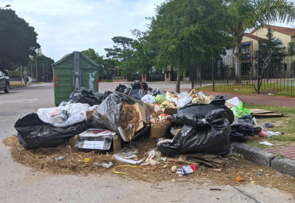 Bolsas de basura y residuos acumulados en el suelo junto a un contenedor verde en un barrio de Montevideo.