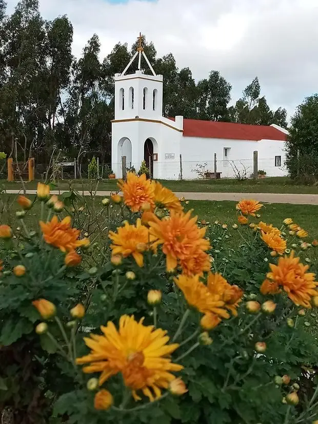 Capilla de Pueblo Edén, escenario de la Fiesta del Churrinche, rodeada de flores naranjas y vegetación serrana. Imagen del sitio puebloeden.com