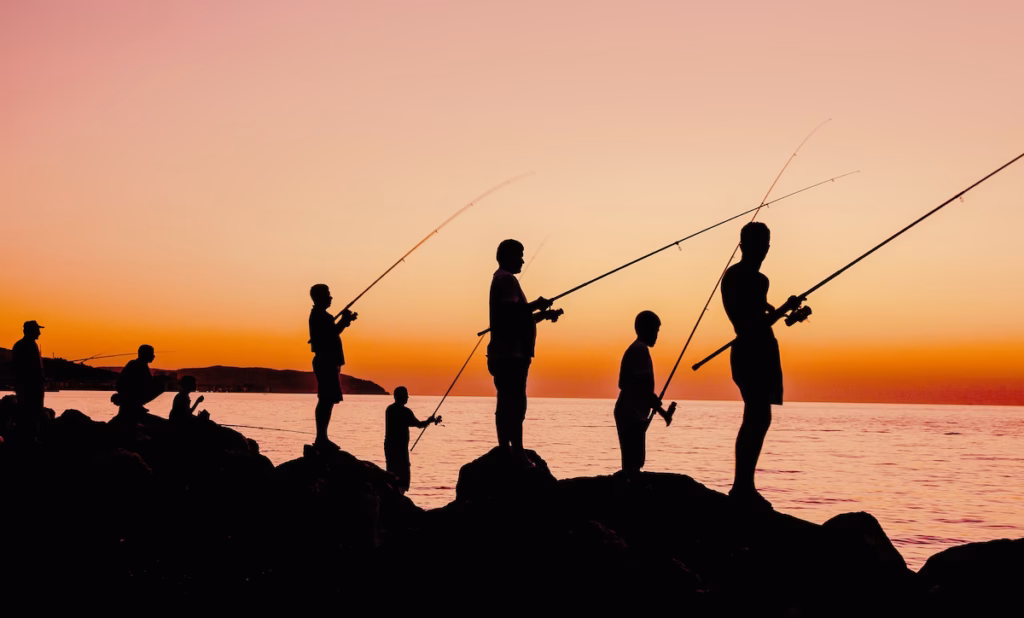 Siluetas de varios pescadores recreativos con cañas en una escollera frente al mar durante el atardecer, representando la pesca deportiva marina en Uruguay.