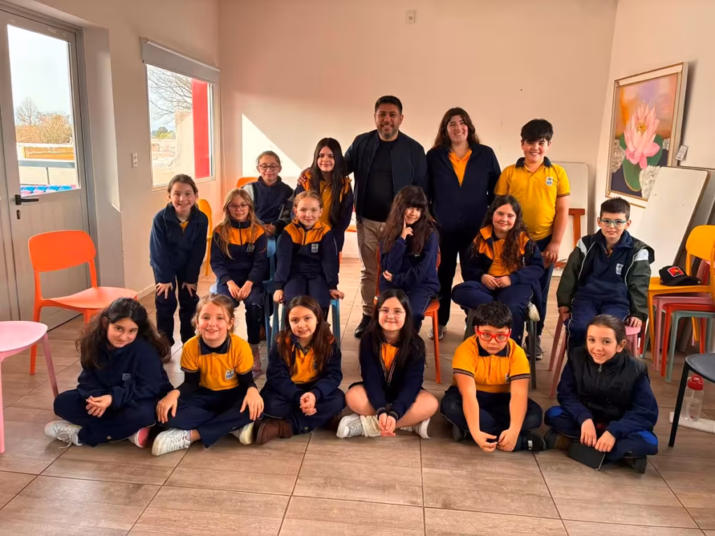 Group of smiling students in navy and yellow uniforms posing with two adults in a bright classroom with colorful chairs and drawings on the wall.